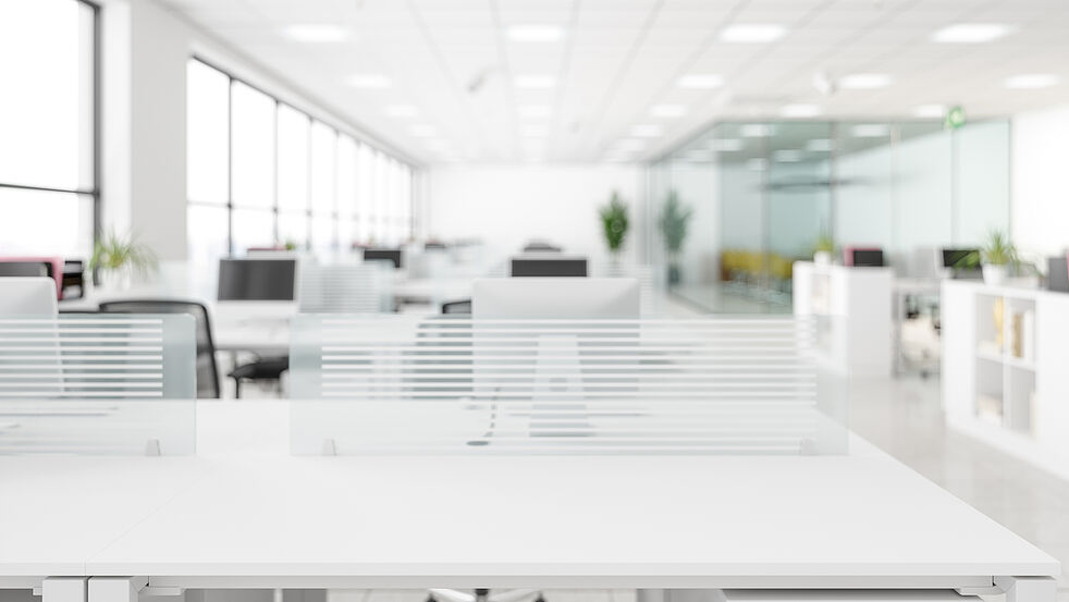Helles, modernes Großraumbüro mit weißen Schreibtischen, abgetrennt durch Glaswände mit dezenten Streifenmustern. Im Hintergrund sind Arbeitsplätze mit Monitoren sowie eine verglaste Besprechungszone zu sehen, große Fenster lassen viel Tageslicht in den Raum.