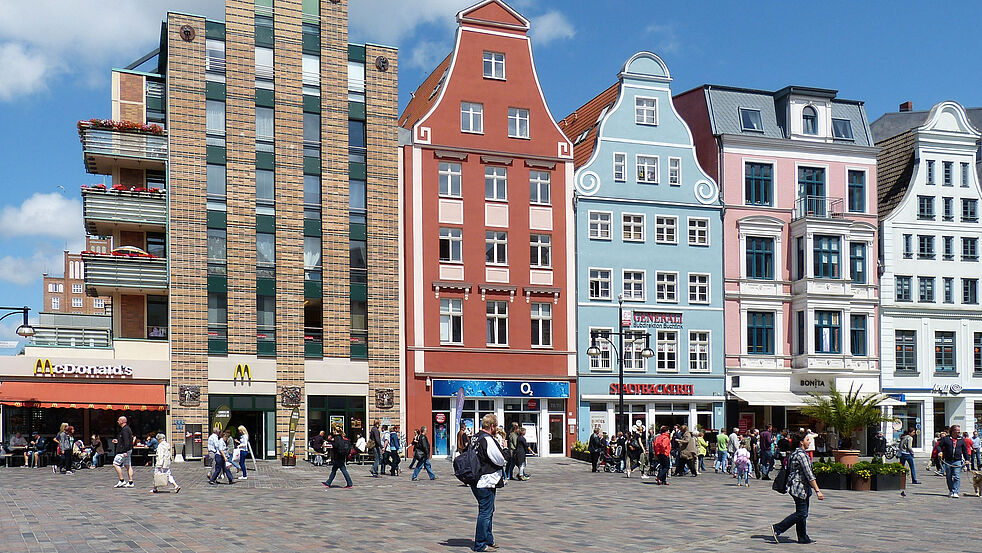 Blick auf die Rostocker Innenstadt mit historischen und modernen Fassaden, im Erdgeschoss zahlreiche Geschäfte und Passanten auf dem belebten Platz.