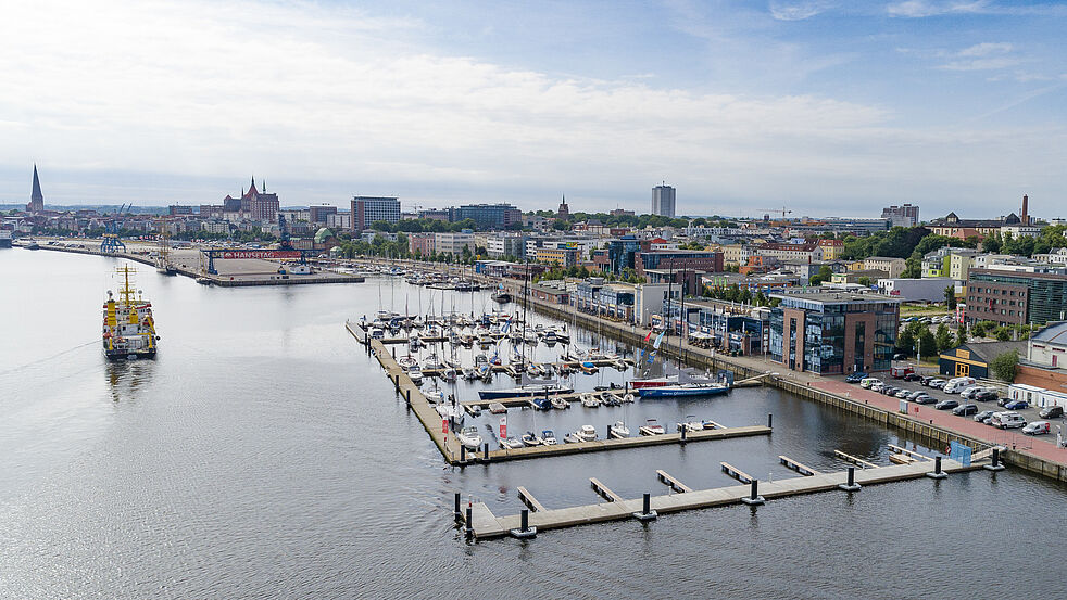 Luftaufnahme des Stadthafens Rostock mit Yachthafen, Gewerbegebäuden an der Kaikante und Blick auf die Innenstadt mit markanten Kirchtürmen und dem Rathaus im Hintergrund.