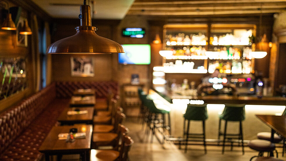 Gemütliche Bar mit warmem Licht, ledergepolsterter Sitzbank und Holztischen auf der linken Seite. Im Hintergrund leuchtet ein gut bestücktes Regal hinter der Theke, vor der mehrere grüne Barhocker stehen. Die Atmosphäre wirkt einladend und entspannt.