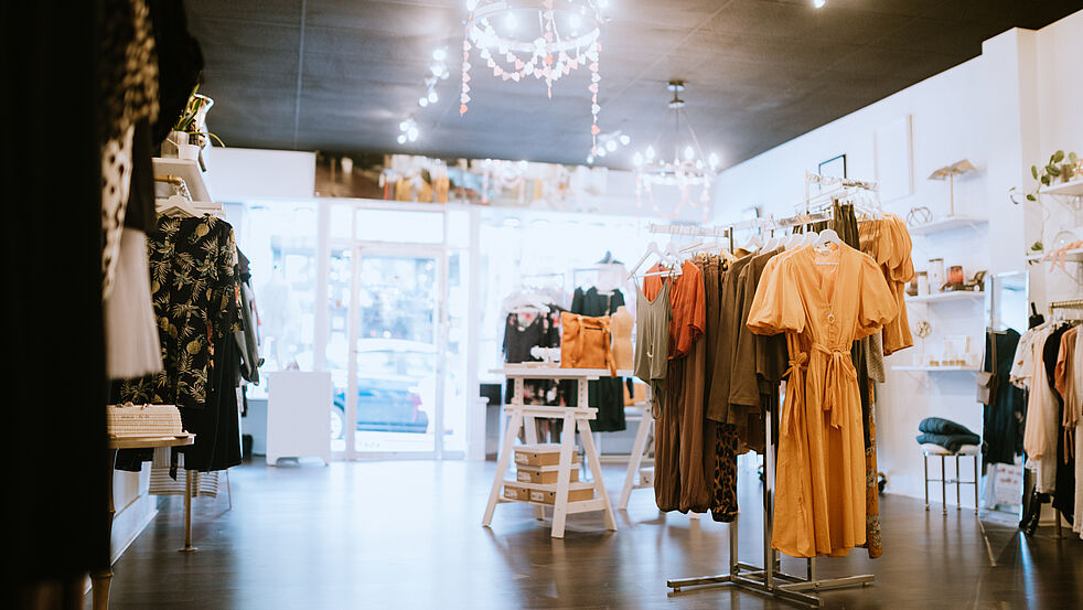 Einladender Modeboutique-Innenraum mit dekorativ beleuchtetem Verkaufsraum. Zwischen zarten Deckenleuchten hängen Kleiderständer mit farblich abgestimmter Kleidung, im Vordergrund ein senfgelbes Kleid. Helle Wände, Accessoires und Pflanzen verleihen dem Laden eine moderne und gemütliche Atmosphäre.